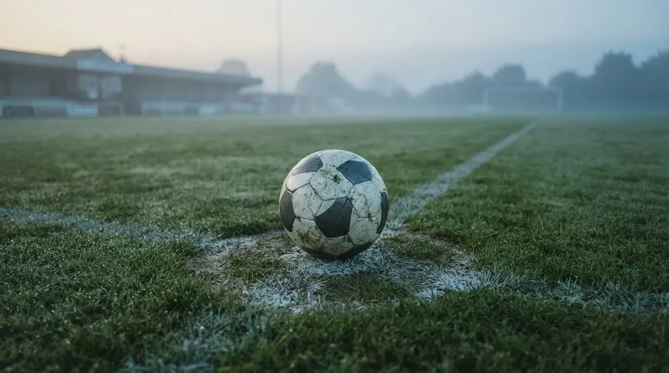 Ein einzelner Fussball liegt auf dem Anstosspunkt eines leeren Rasenplatzes am frühen Morgen, Tau glitzert auf dem Gras, die Tribüne liegt im Dunst.