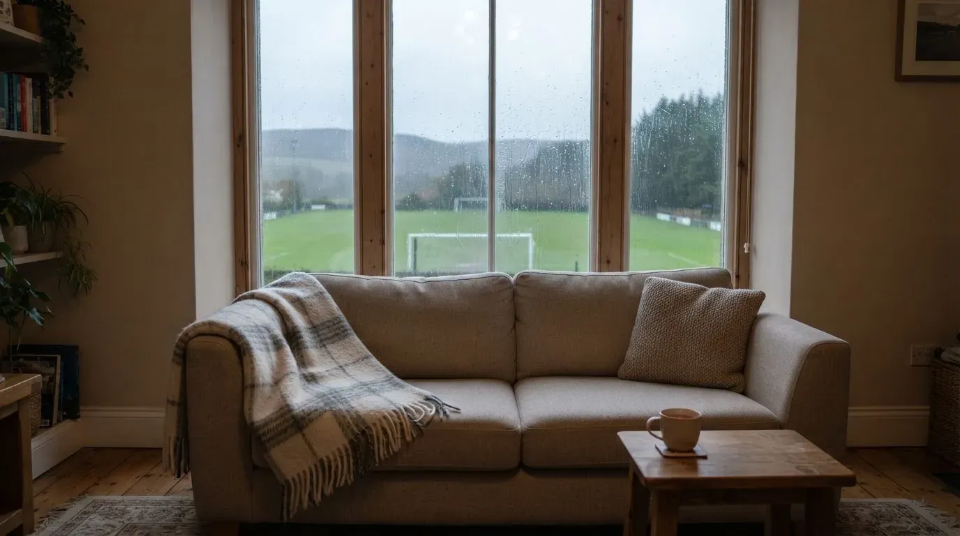 Ein Sofa mit Wolldecke steht vor einem grossen Fenster, draussen Regen und in der Ferne ein Fussballplatz.