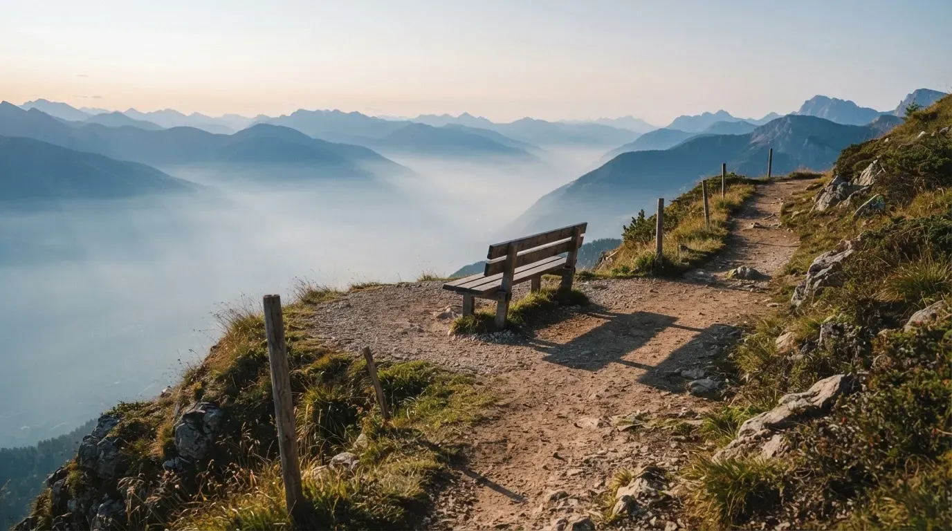 Ein Wanderweg führt an einem Aussichtspunkt über einem Bergtal im Morgennebel vorbei, am Wegrand steht eine einfache Holzbank.