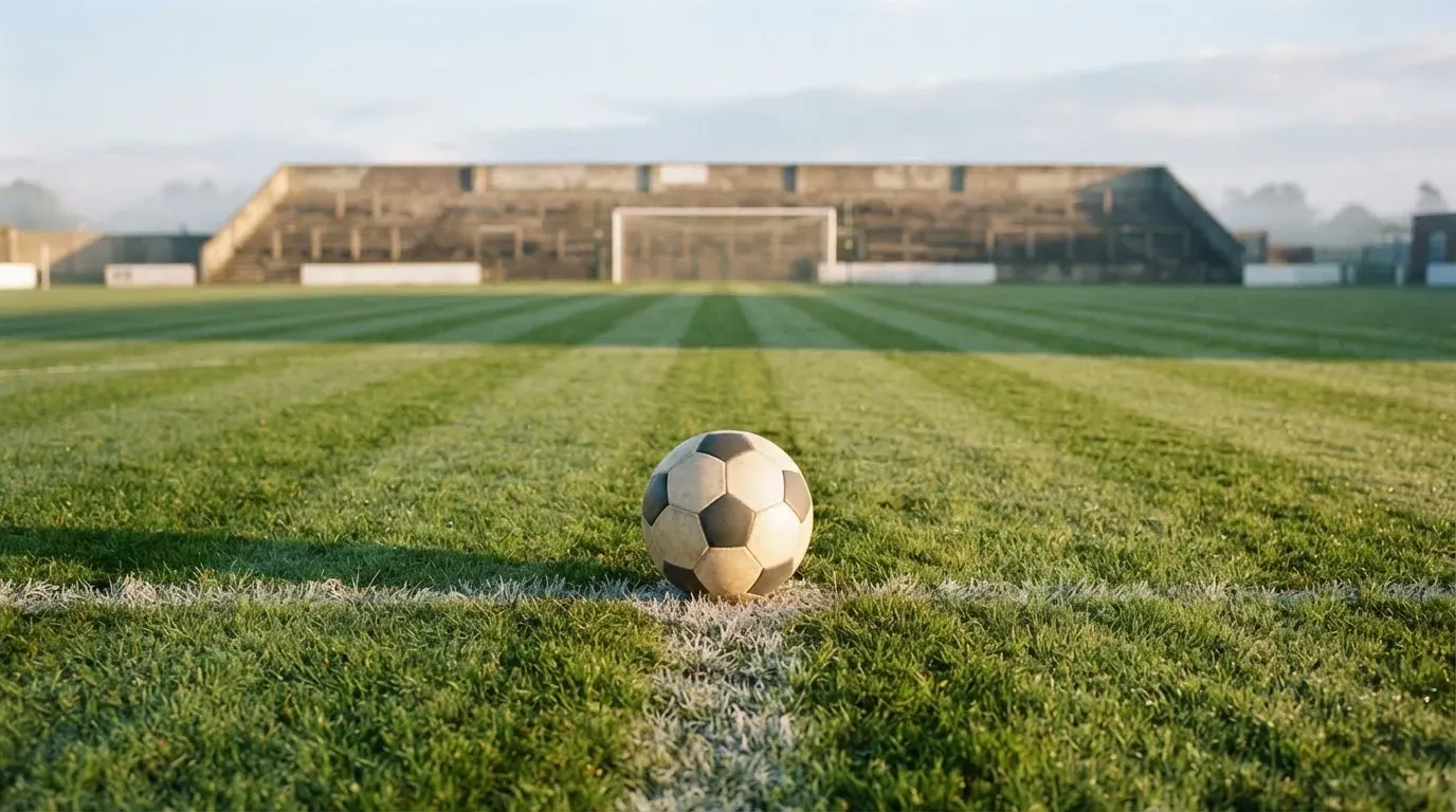 Ein Fussball liegt auf dem Anstosspunkt eines leeren Rasenplatzes im frühen Morgenlicht, dahinter erhebt sich eine menschenleere Tribüne.