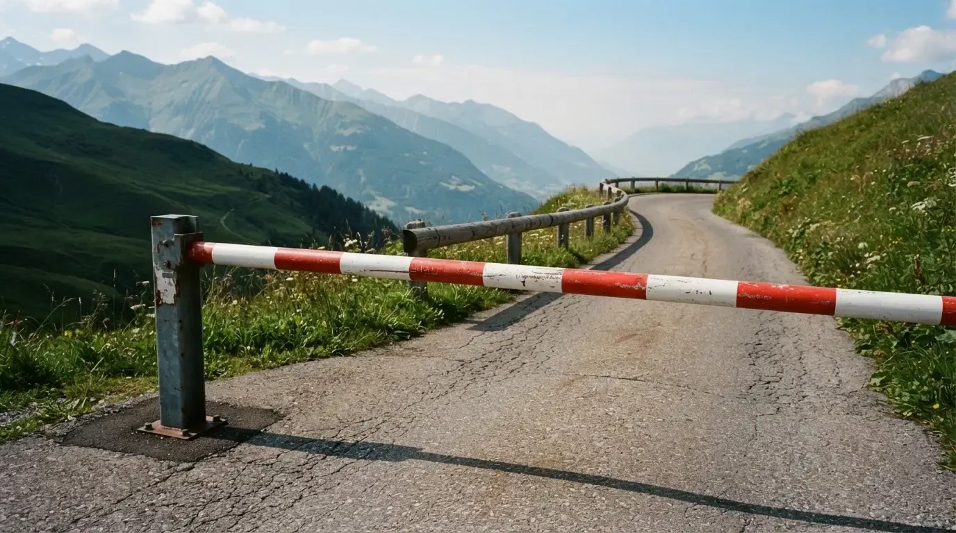 Ein Schlagbaum sperrt eine Landstrasse in den Bergen, dahinter windet sich der Weg zwischen grünen Hängen.