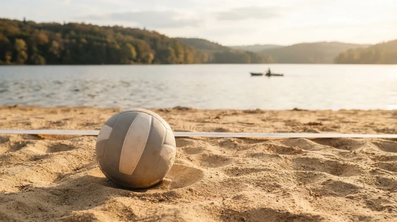 Ein Volleyball liegt im Sand eines Spielfelds am Seeufer, im Hintergrund gleitet ein Boot über das Wasser.