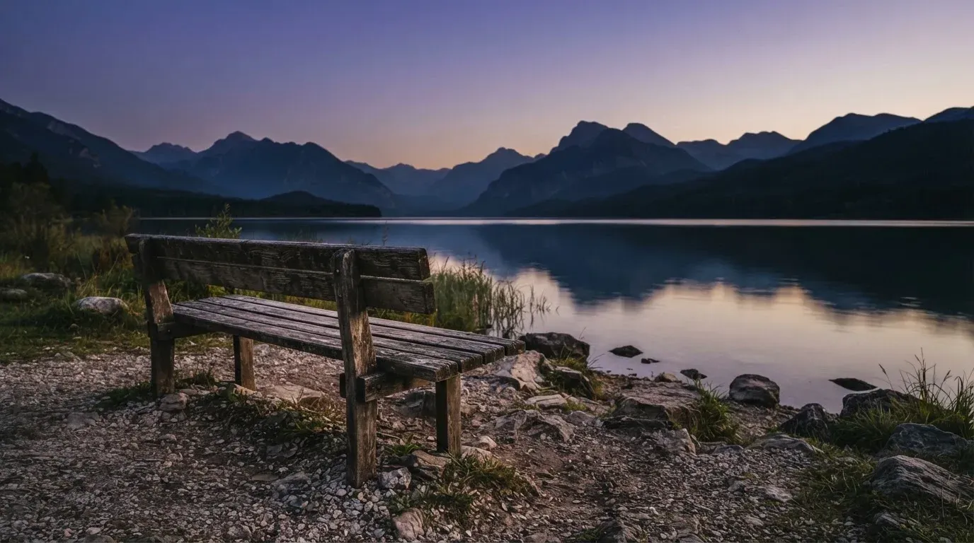 Eine Holzbank steht in der Dämmerung am Seeufer, im Hintergrund zeichnet sich ein Bergpanorama gegen den violetten Himmel ab.