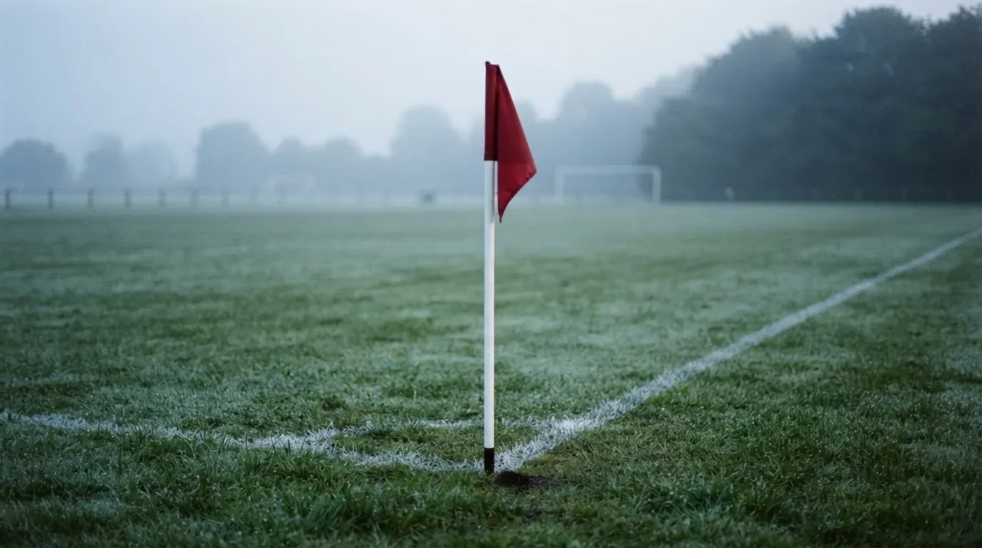 Eine Eckfahne steht am Morgen auf nassem Rasen, Tautropfen hängen im Gras und über dem Platz liegt leichter Dunst.
