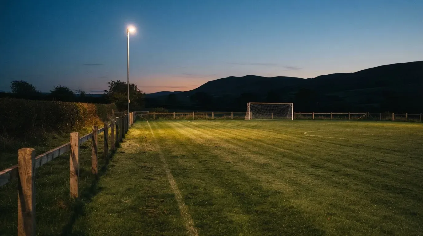 Ein kleiner Dorf-Fussballplatz liegt in der Dämmerung unter einem hohen Flutlichtmast, am Spielfeldrand steht ein Holzzaun.