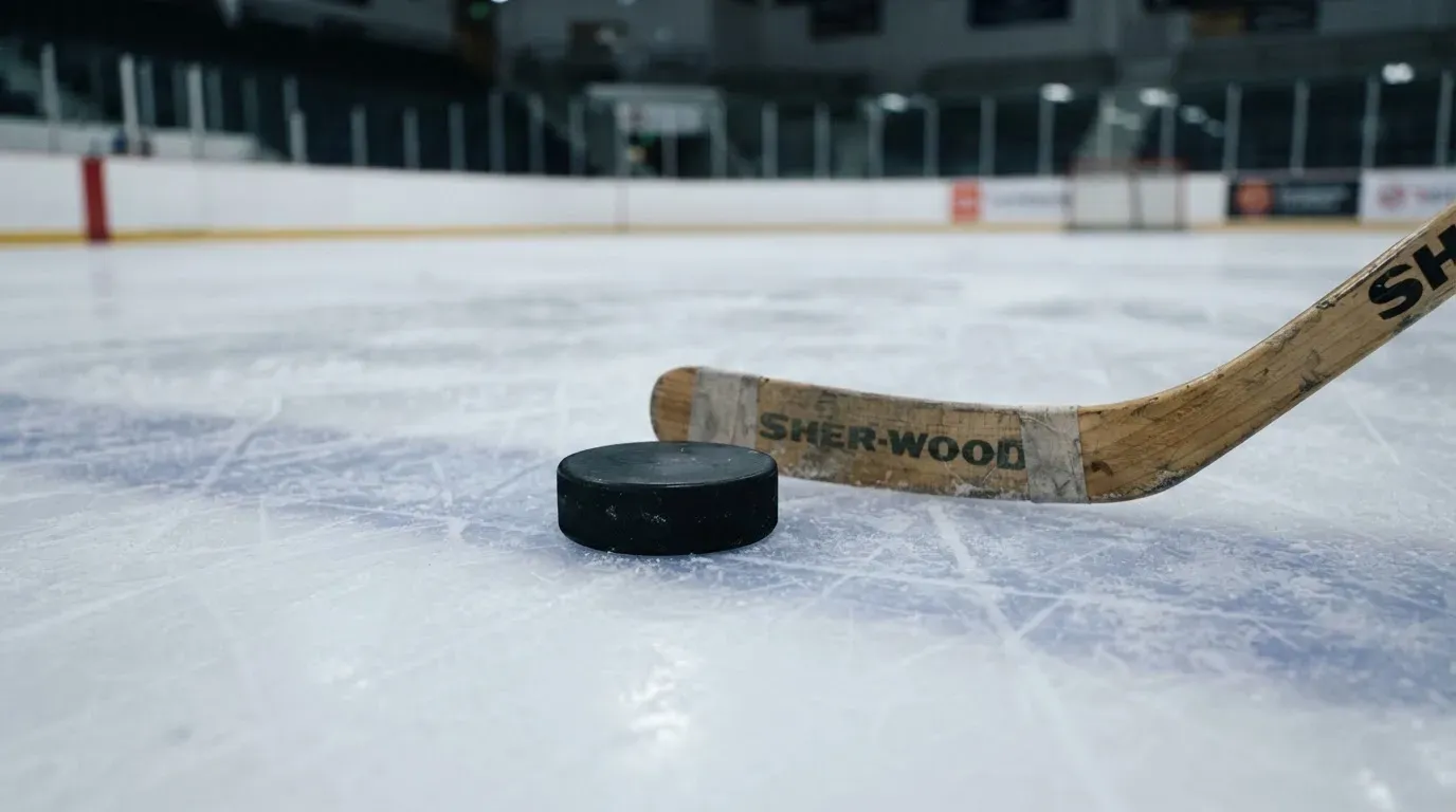 Ein Eishockeypuck liegt auf der Eisfläche neben dem Blatt eines Schlägers, die Bande dahinter ist im Dunkeln nur schwach zu erkennen.