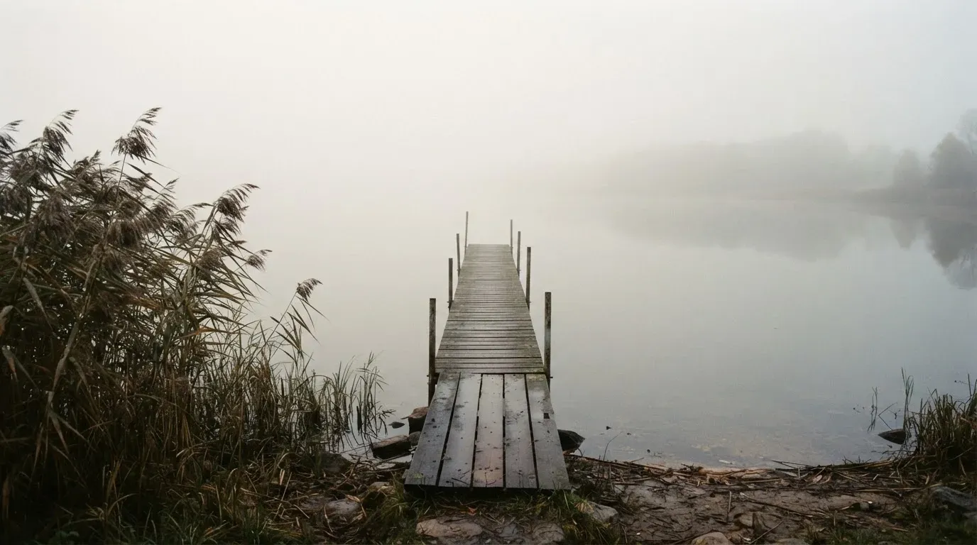 Ein Seeufer mit Schilf und einem schmalen Holzsteg liegt im Nebel, das Wasser ist ruhig und das andere Ufer kaum sichtbar.
