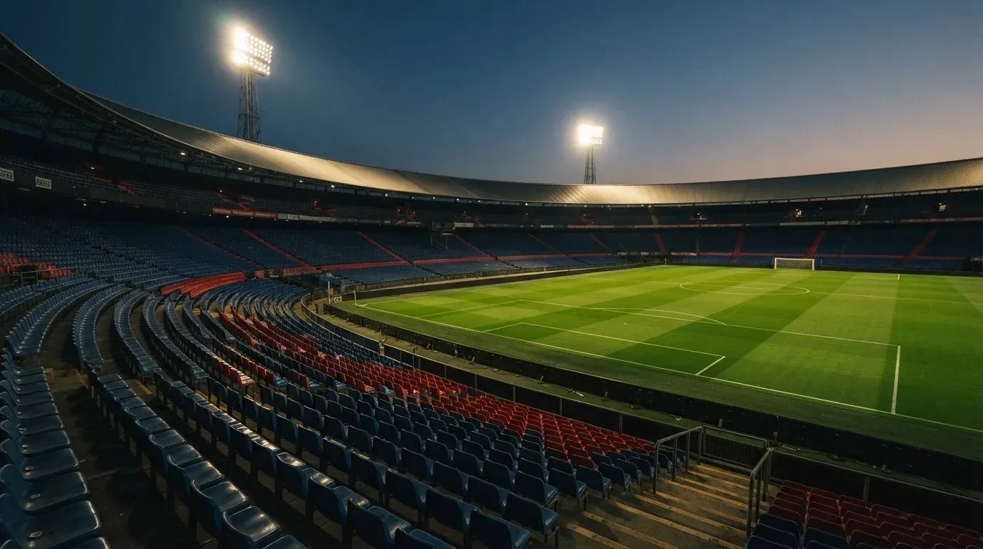 Leere Tribünenränge eines Fussballstadions in der Abenddämmerung, über dem Rasen leuchten die Flutlichter.