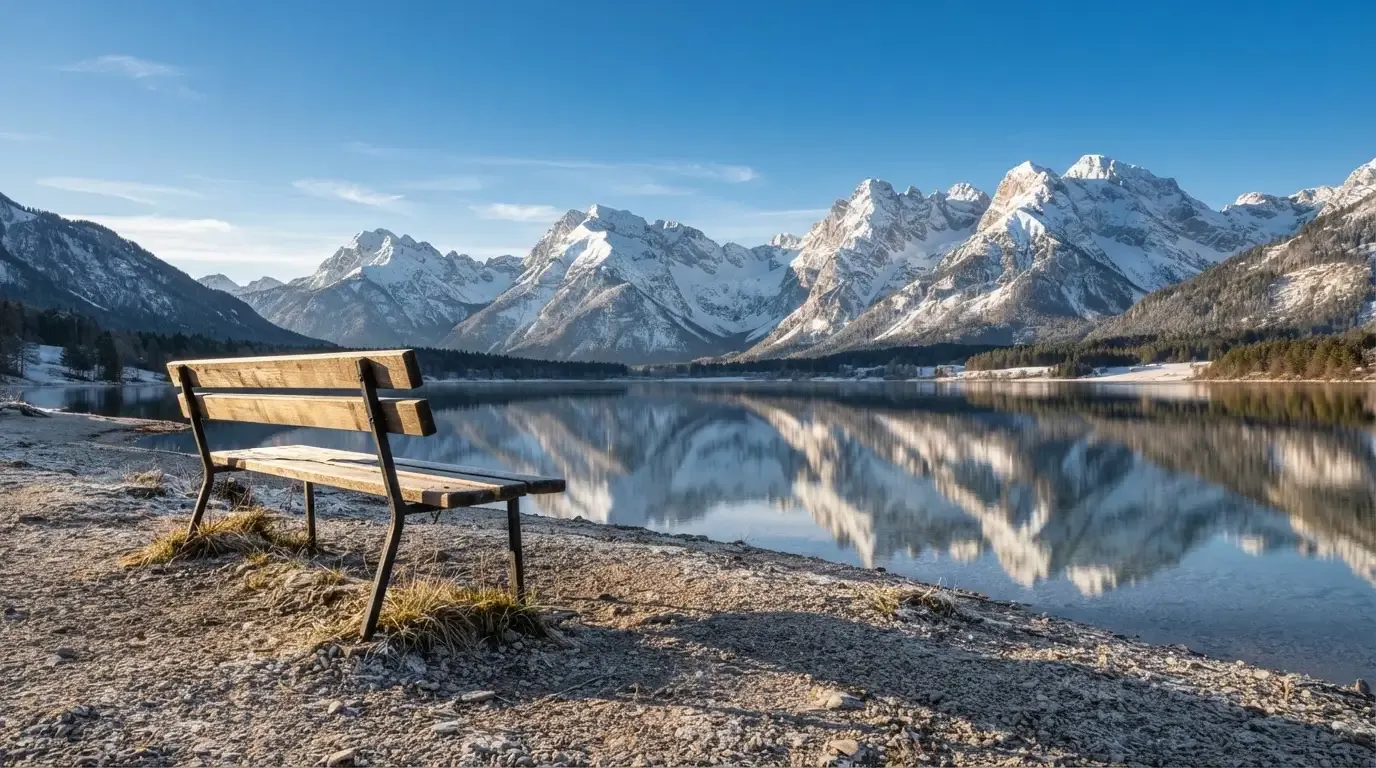 Eine Bank an einem Bergsee, dahinter schneebedeckte Gipfel im klaren Licht eines Wintermorgens.