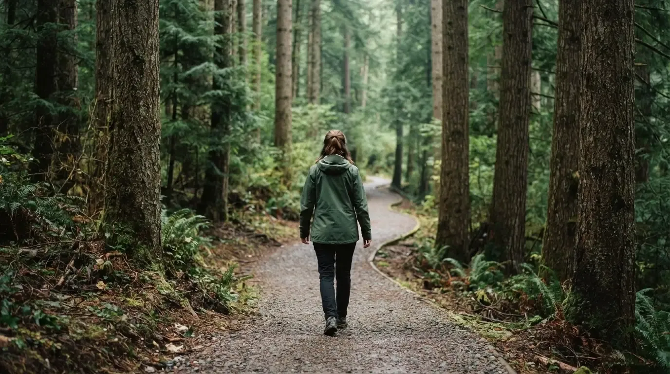 Eine Person von hinten auf einem Waldweg, die langsam zwischen hohen Tannen geht.