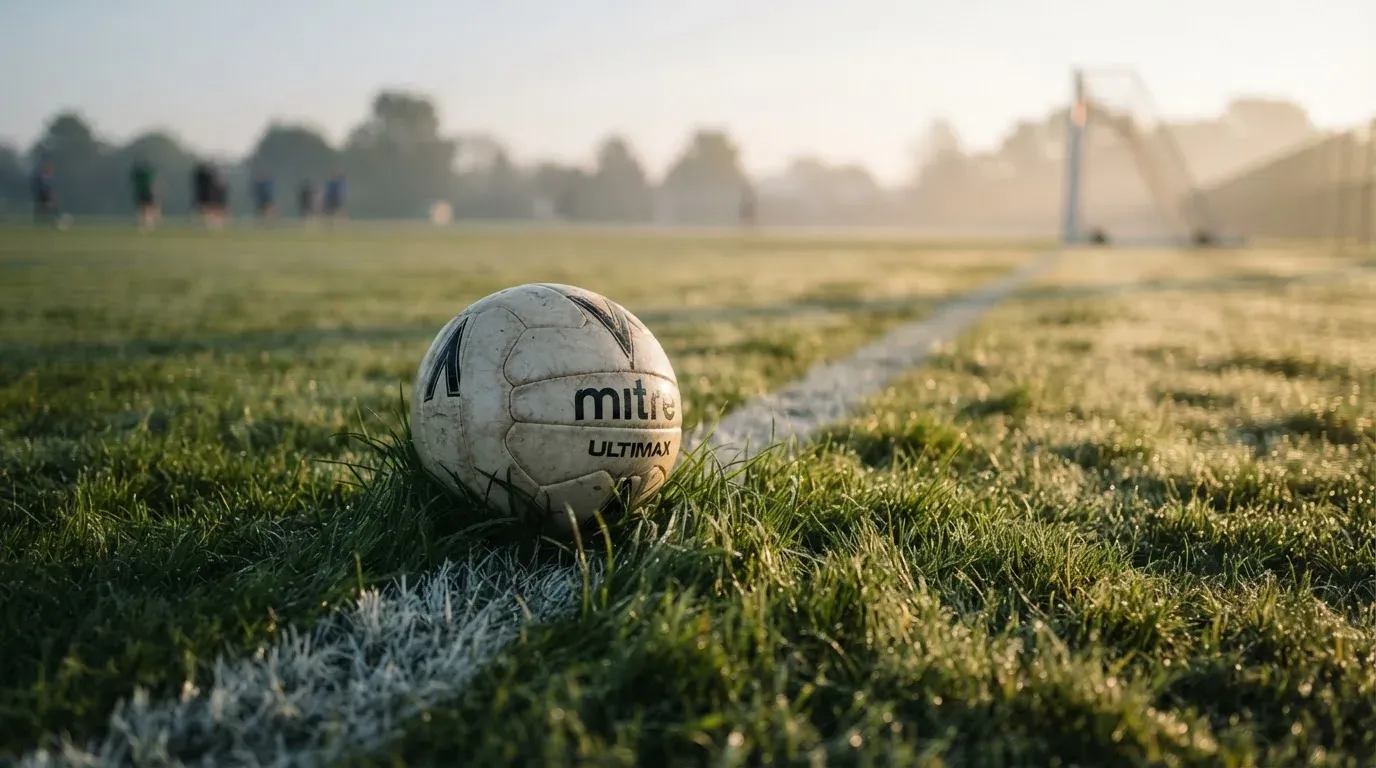 Ein Fussball ruht an der Seitenlinie im taufeuchten Gras, die ersten Sonnenstrahlen fallen schräg über den Platz.