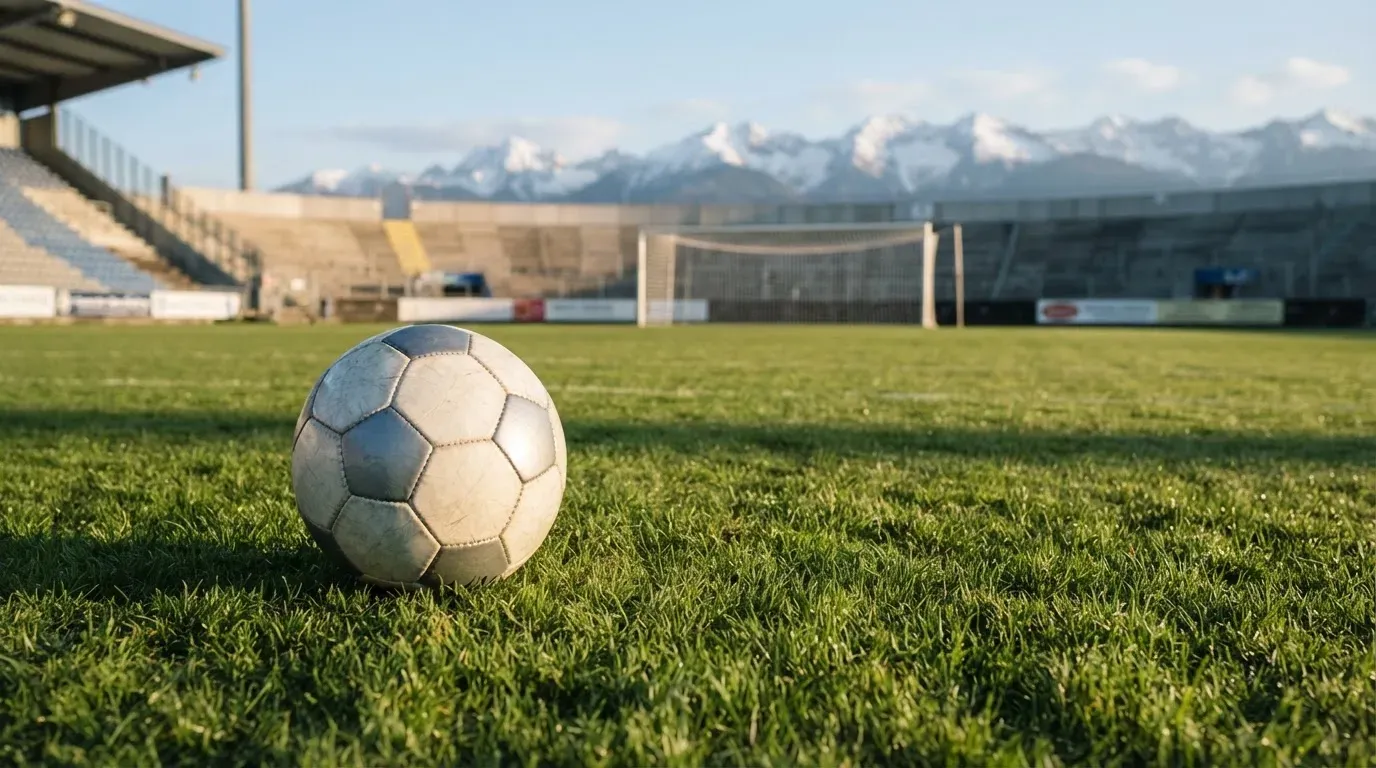 Ein Fussball liegt auf dem Rasen eines leeren Stadions am frühen Abend, dahinter erheben sich schneebedeckte Berge unter klarem Himmel.