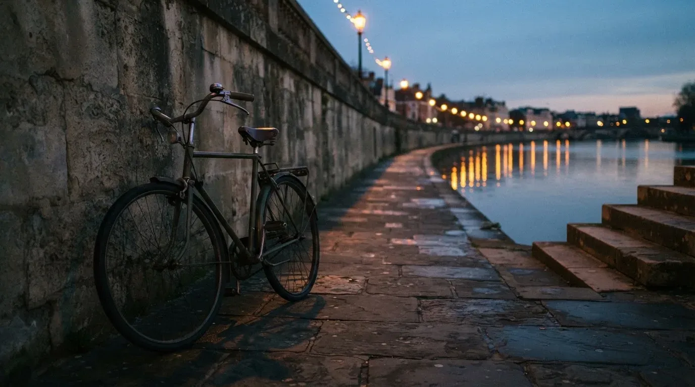 Ein Velo lehnt an einer Steinmauer am Ufer eines Flusses, die Strassenlaternen leuchten in der Dämmerung.
