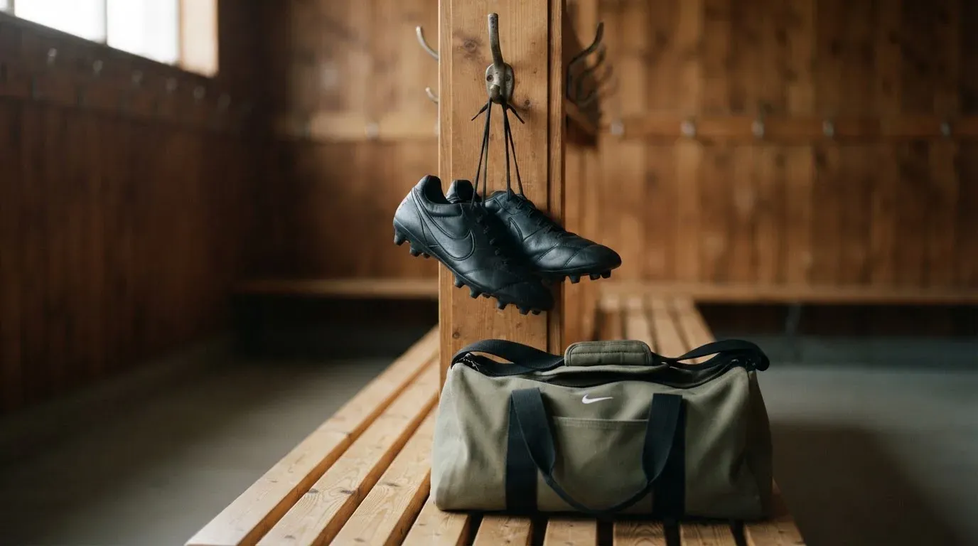Ein Paar Fussballschuhe hängt an einem Haken in einer ruhigen Umkleide, darunter steht eine Sporttasche auf einer Holzbank.