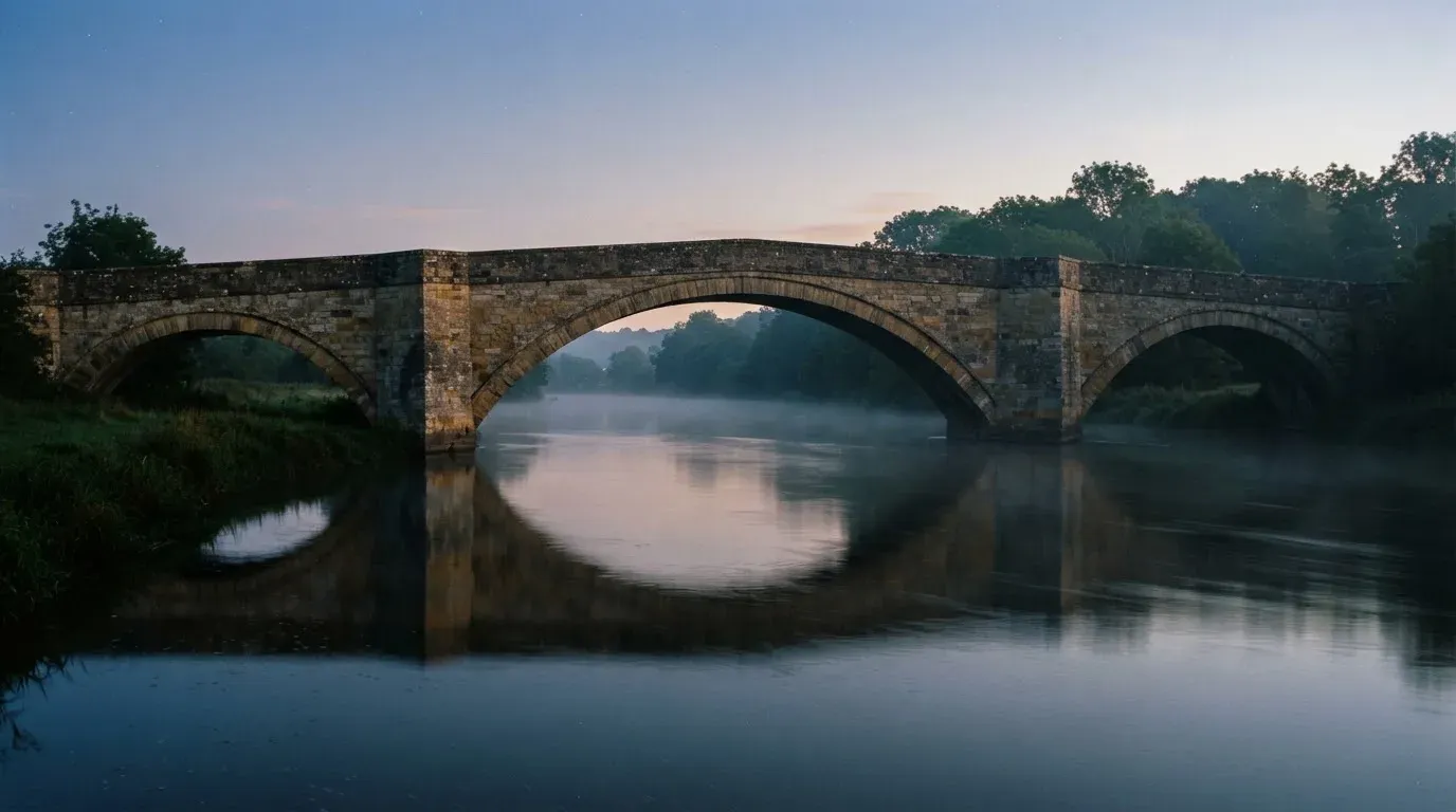 Eine Steinbrücke spannt sich in der Dämmerung über einen Fluss, auf dem Wasser liegt leichter Nebel.