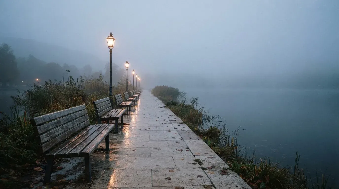 Eine Seepromenade mit leeren Bänken an einem nebligen Morgen, im Wasser spiegeln sich die Lichter der Uferlaternen.