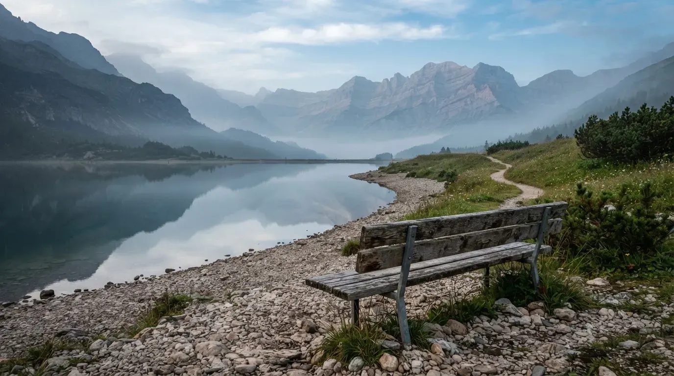 Eine Holzbank an einem Seeufer mit Blick auf eine Bergkette im Morgendunst, das Wasser ist spiegelglatt.