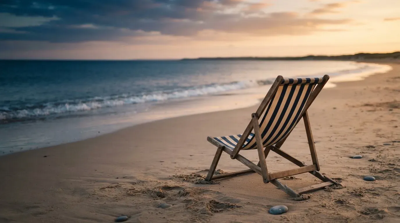 Ein leerer Liegestuhl aus Holz steht am Rand eines Sandstrands, Abendlicht und ruhige Wellen im Hintergrund.