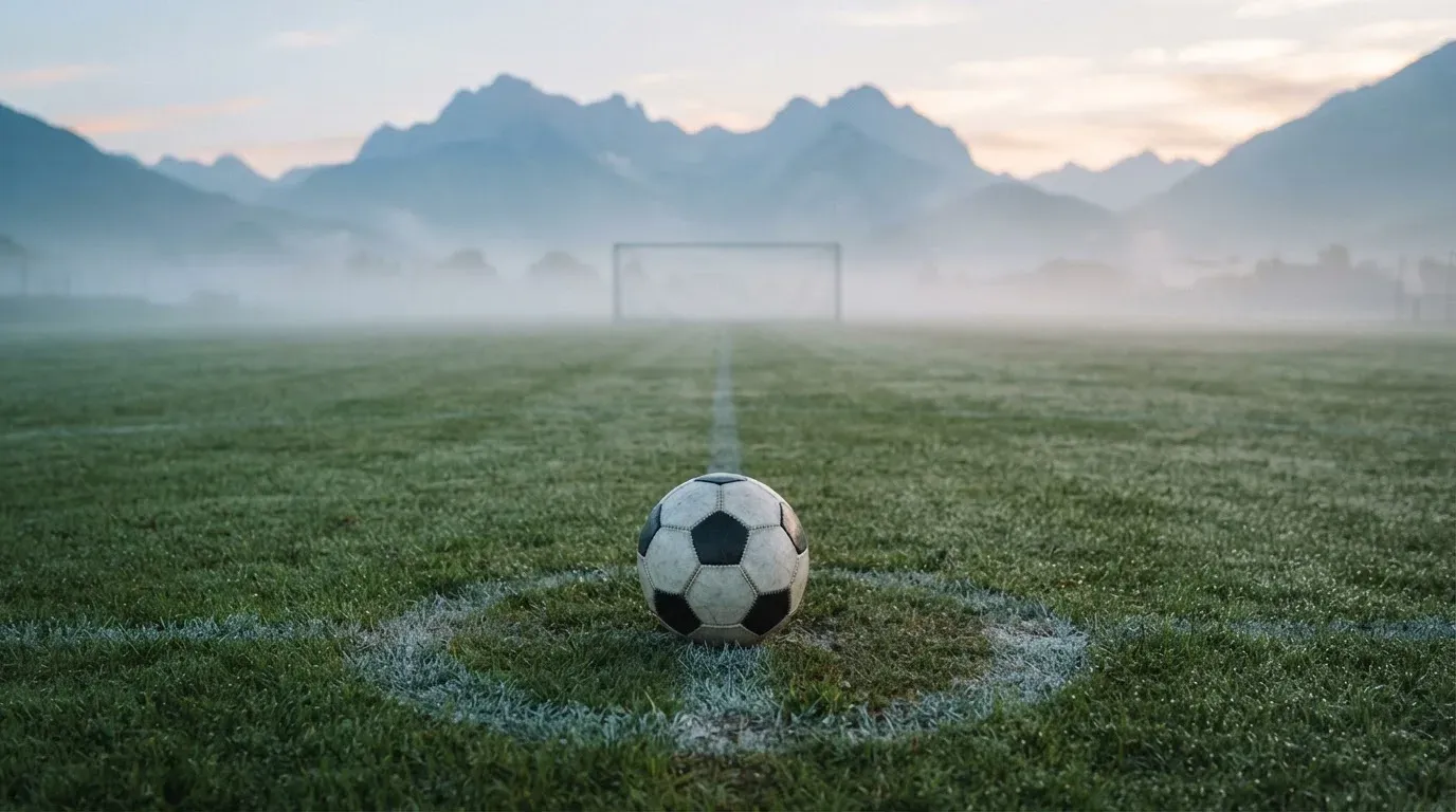 Ein einzelner Fussball auf dem Mittelpunkt eines leeren Rasens im Morgennebel, hinter dem Tor ragen Berge auf