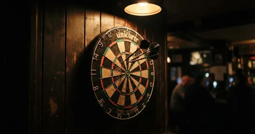 Drei Dartpfeile stecken in einer Dartscheibe an einer Holzwand in einer dämmrigen Kneipe, warmes Lampenlicht fällt von oben