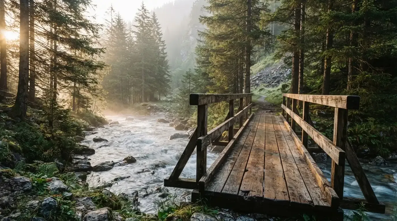Eine alte Holzbrücke führt über einen Bergbach im Morgenlicht, Tannen säumen das Ufer