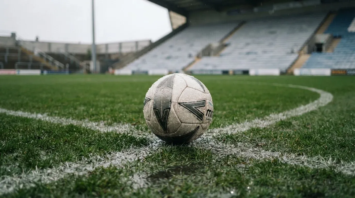 Ein Fussball liegt auf dem Anstosspunkt eines nassen Rasens, dahinter eine leere Tribüne unter grauem Himmel.