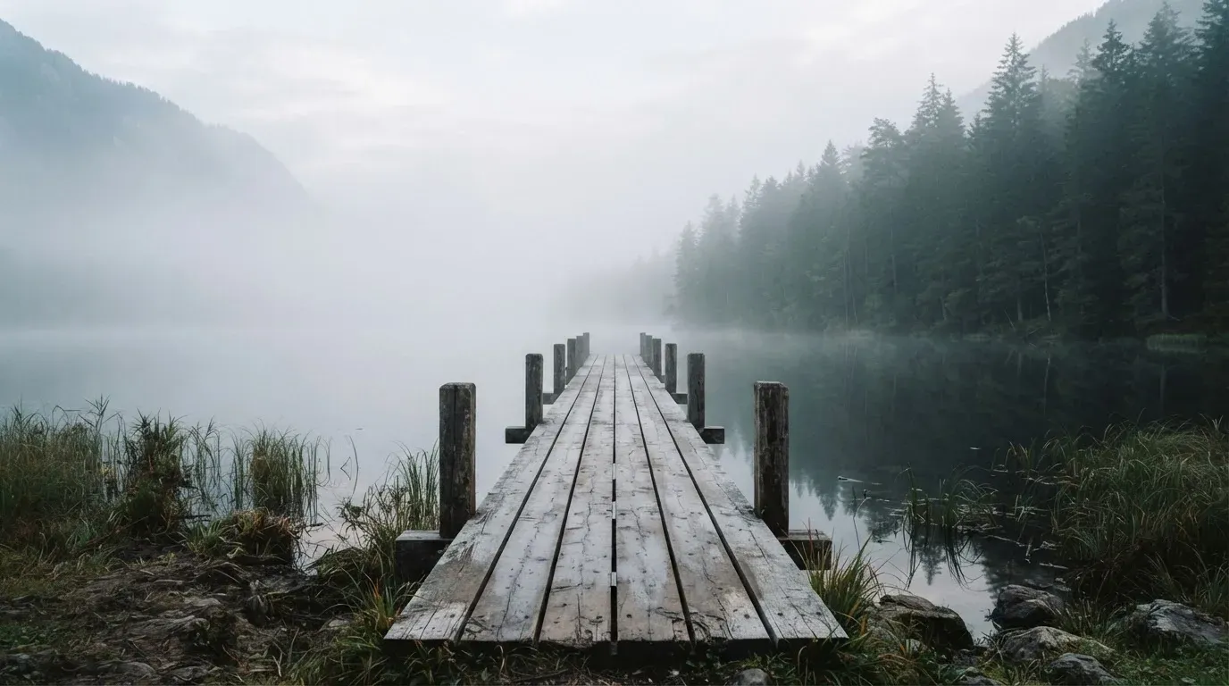 Ein Holzsteg führt über einen ruhigen Bergsee im Nebel, am anderen Ufer stehen dunkle Tannen.