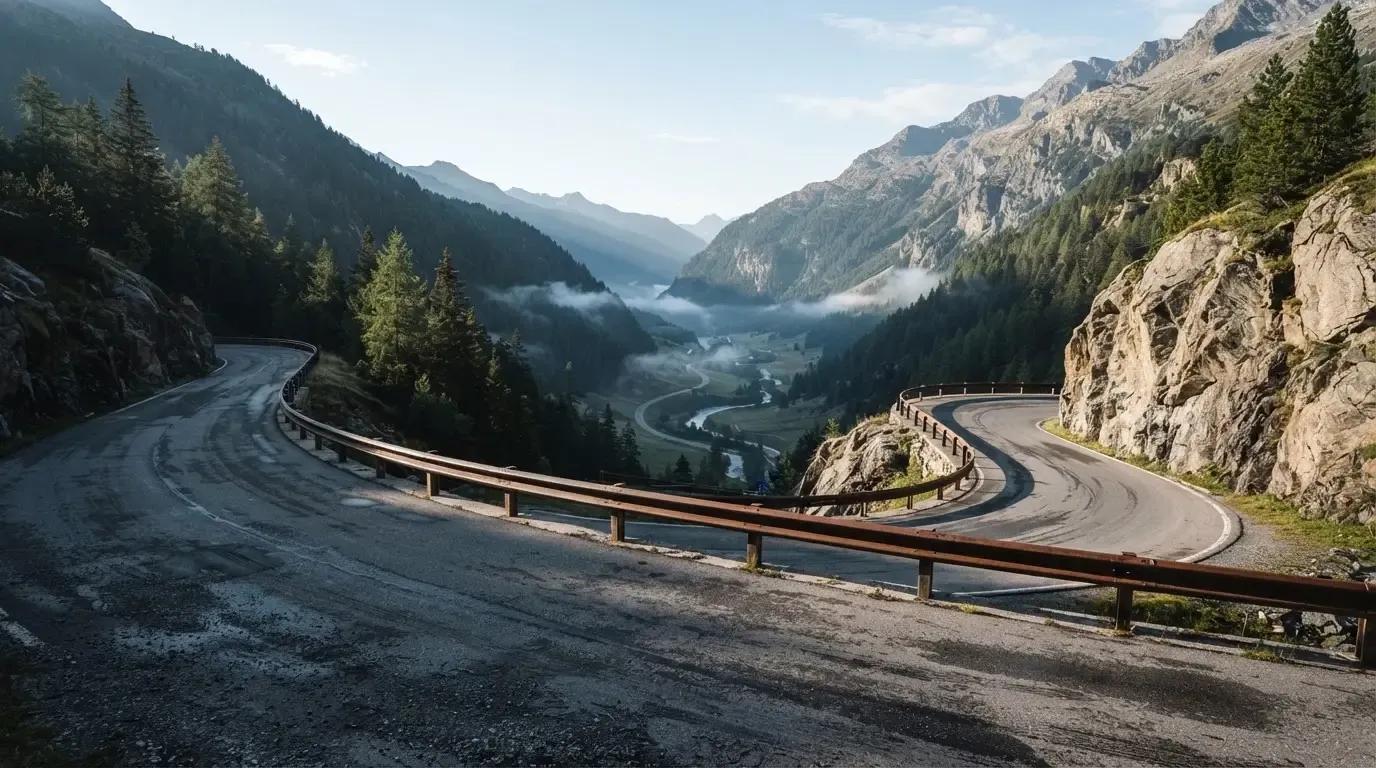 Eine Passstrasse in den Alpen mit enger Kurve und Leitplanke, darüber ein klarer Morgenhimmel und einzelne Nebelschwaden im Tal.