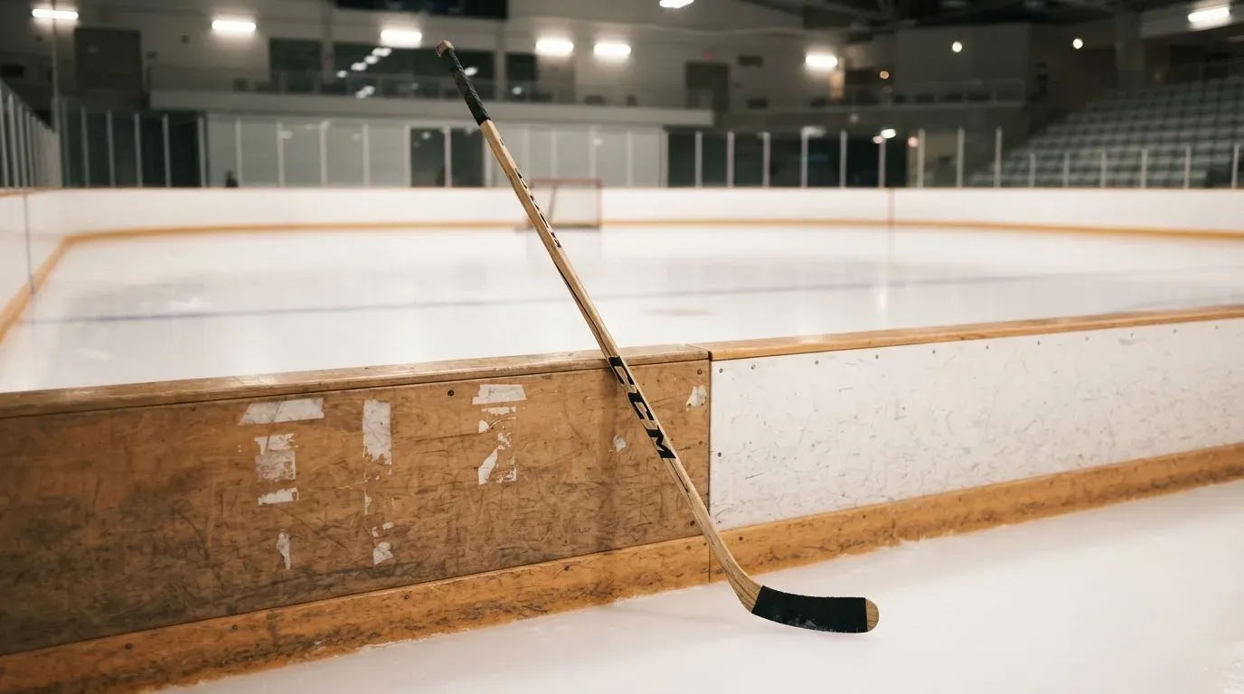 Ein Eishockeyschläger lehnt an der Bande, in der Ferne liegt die hell beleuchtete Eisfläche einer Halle unter Flutlicht.