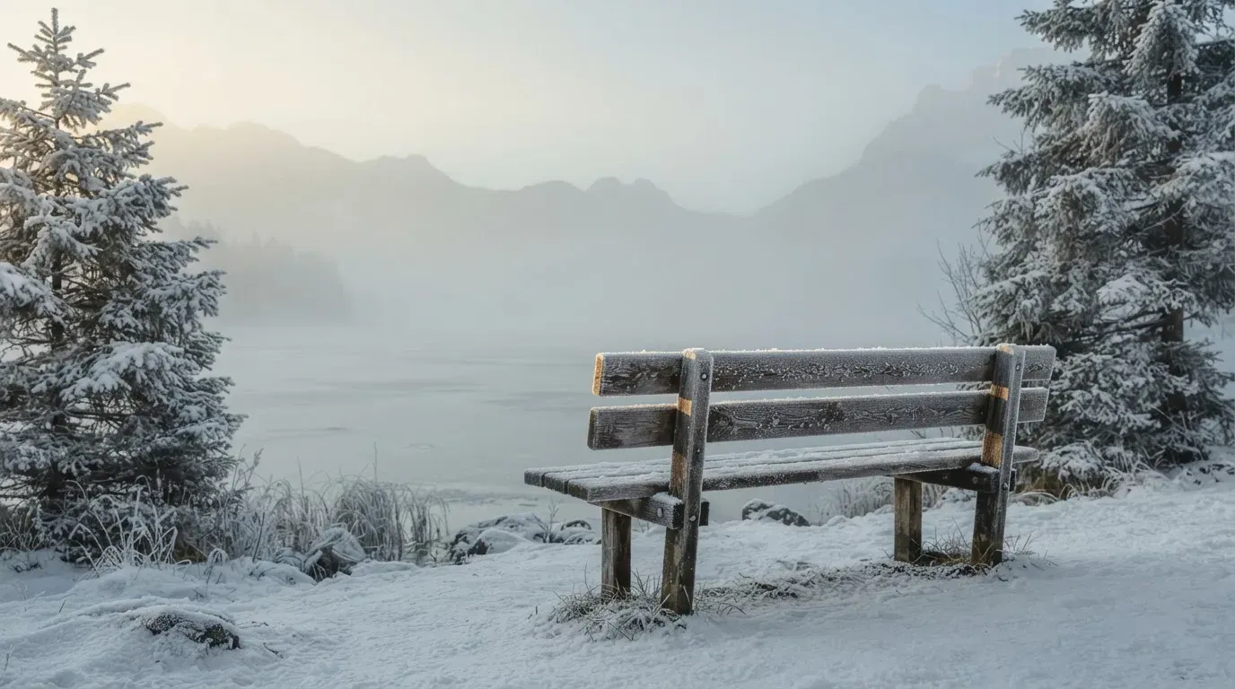 Ein zugefrorener Bergsee im Morgendunst, am Ufer steht eine einsame Holzbank zwischen verschneiten Tannen.