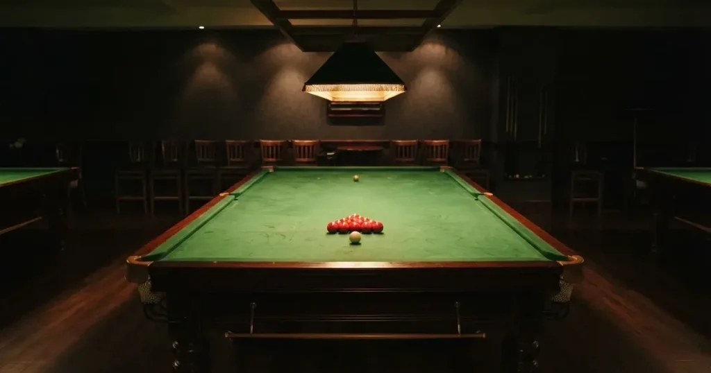 Ein Snookertisch mit grünem Tuch und einem Dreieck aus roten Kugeln unter einer tief hängenden Lampe in einer leeren Halle.