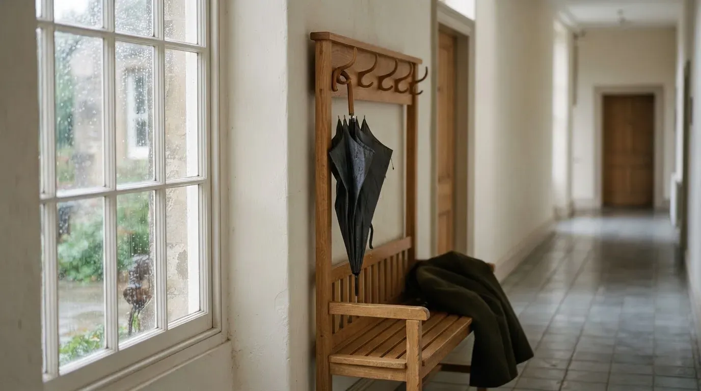 Eine Garderobe aus Holz mit einem dunklen Regenschirm in einem stillen Korridor, am Fenster rinnt Regen herunter.