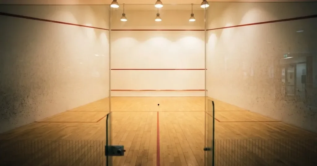 Ein leerer Squashcourt mit weissen Wänden und hellem Holzboden, ein Ball liegt in der Mitte, Deckenlicht spiegelt sich im Parkett.