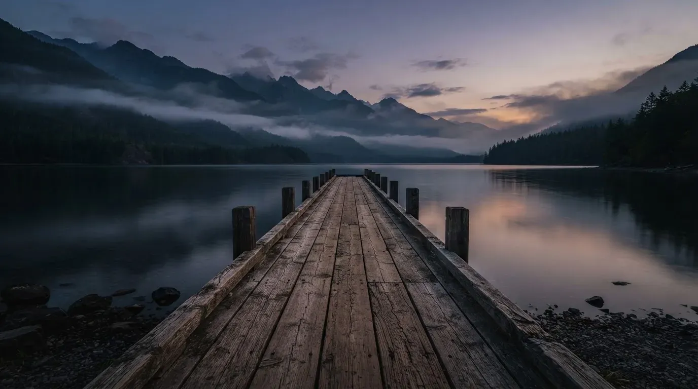 Ein Bergsee in der Dämmerung mit einem leeren Holzsteg, die Wasseroberfläche ist ruhig und Nebel zieht über die Hänge.