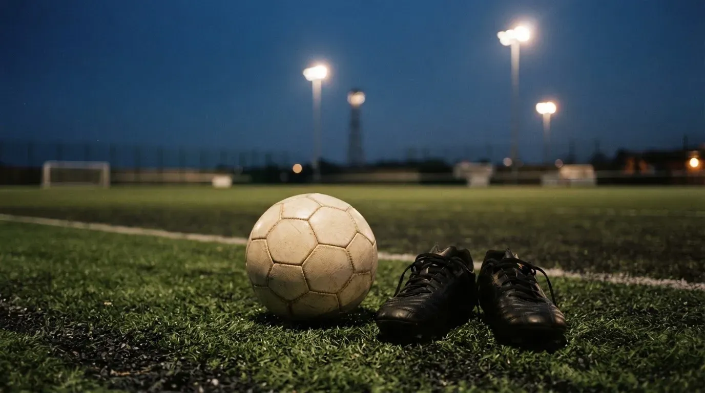Ein Fussball und ein Paar Stollenschuhe stehen am Rand eines Kunstrasenplatzes, Flutlichtmasten ragen in den Abendhimmel.