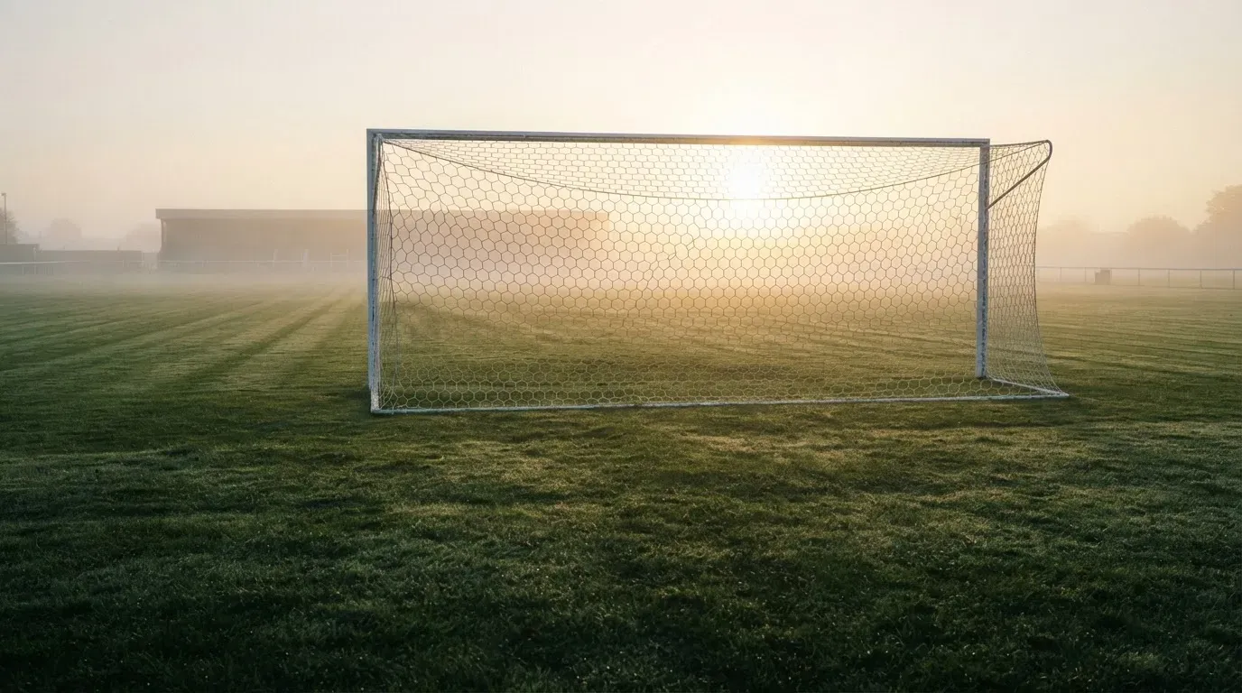 Ein Torpfosten mit Netz steht im Gegenlicht auf einem leeren Fussballplatz, über dem Rasen liegt Nebel.