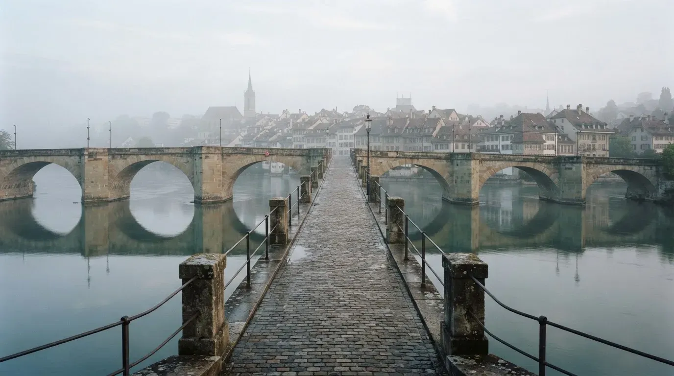 Eine Steinbrücke führt im Frühnebel über einen Fluss, auf dem Fussweg ist niemand zu sehen.