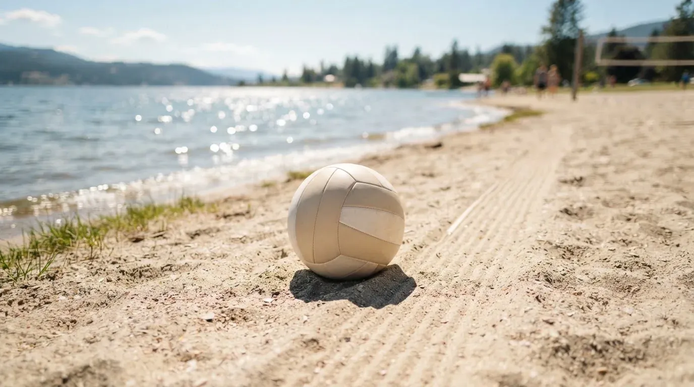 Ein Volleyball liegt auf dem Sand eines Strandfeldes am Seeufer, im Hintergrund glitzert das Wasser.