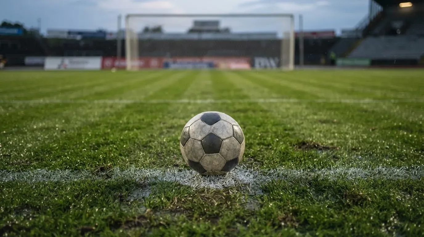Ein einzelner Fussball liegt auf dem Elfmeterpunkt eines leeren Strafraums, der Rasen glänzt nass.