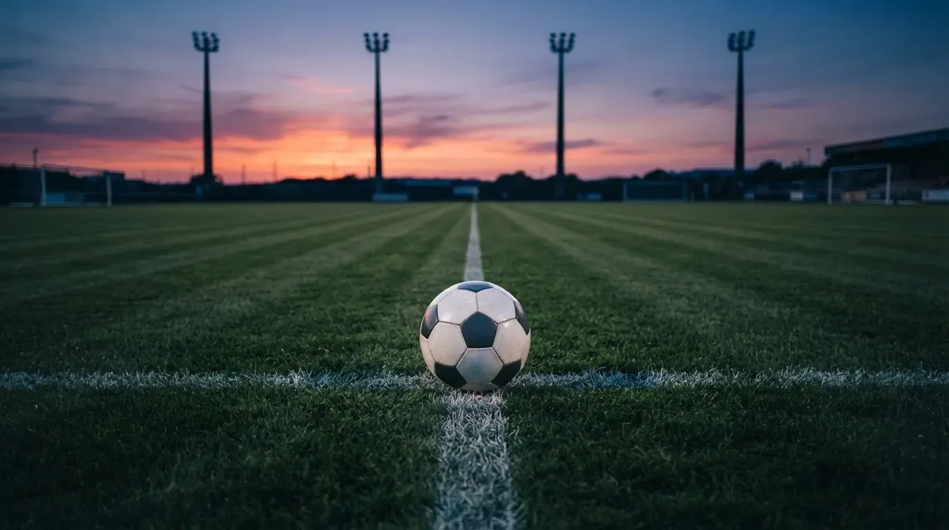 Ein Fussball liegt auf dem Mittelpunkt eines leeren Rasenplatzes, im Hintergrund ragen Flutlichtmasten in die Abenddämmerung