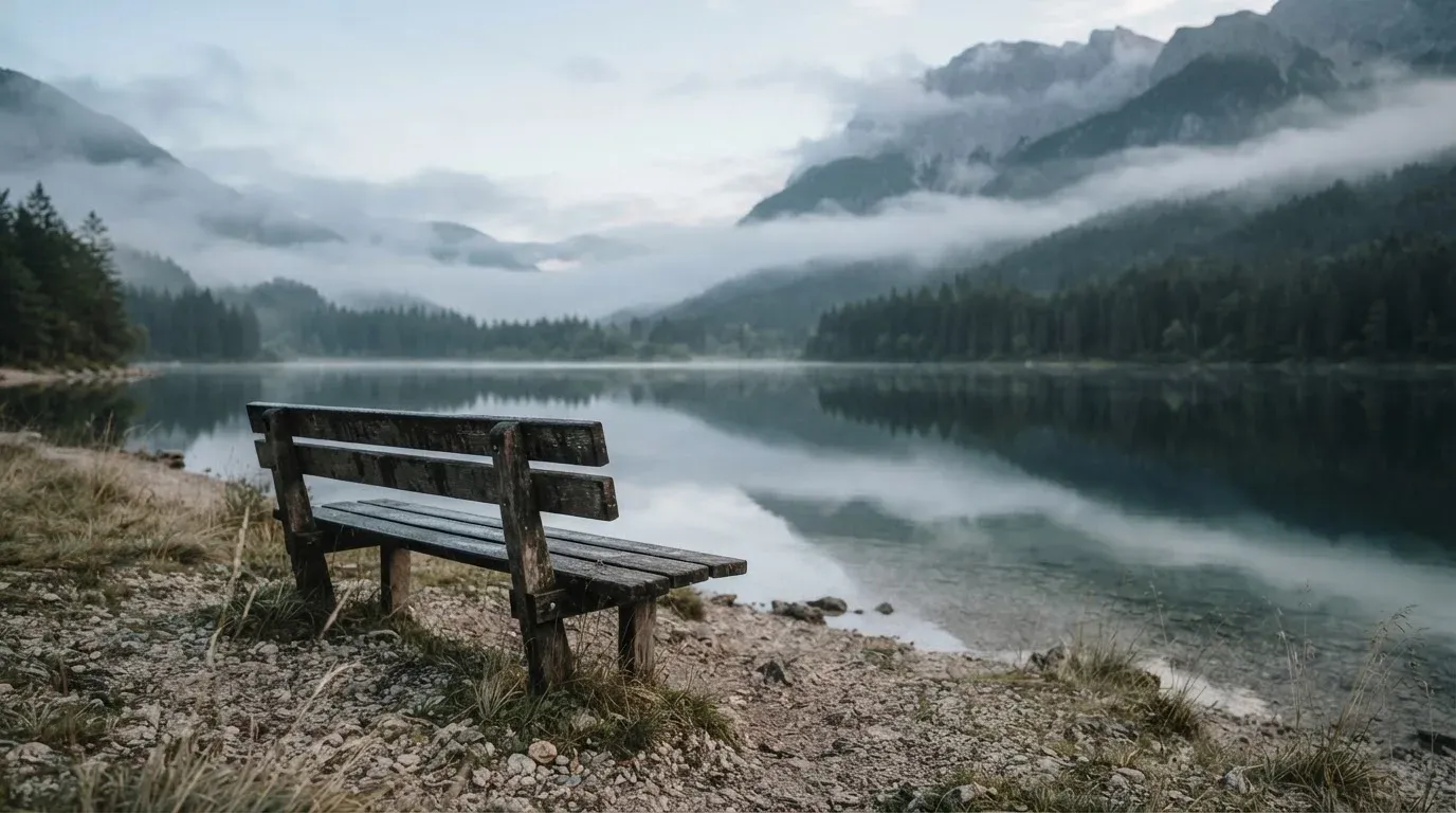 Eine Holzbank am Ufer eines ruhigen Bergsees, am gegenüberliegenden Ufer steigen dunkle Hänge in den Frühnebel