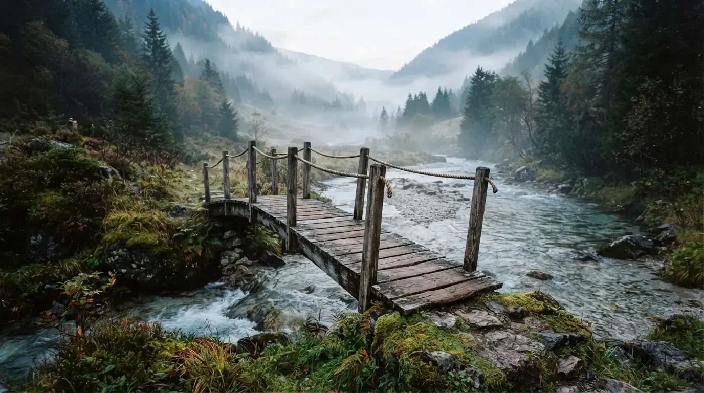 Eine alte Holzbrücke über einen Gebirgsbach, Moos auf den Steinen, Nebelschwaden hängen im Tal
