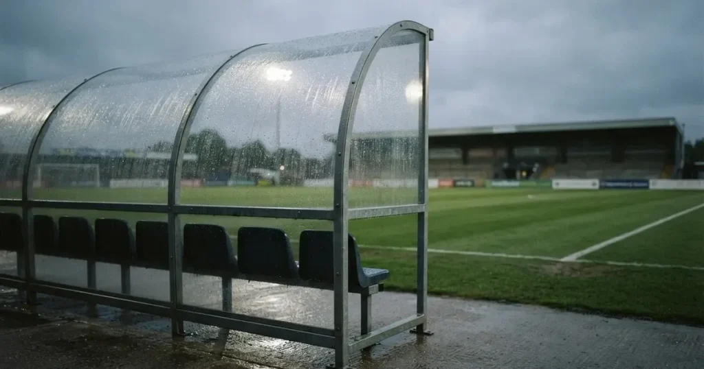 Eine leere Ersatzbank unter einem Plexiglasdach am Spielfeldrand, Regentropfen auf der Scheibe, dahinter ein Rasenplatz