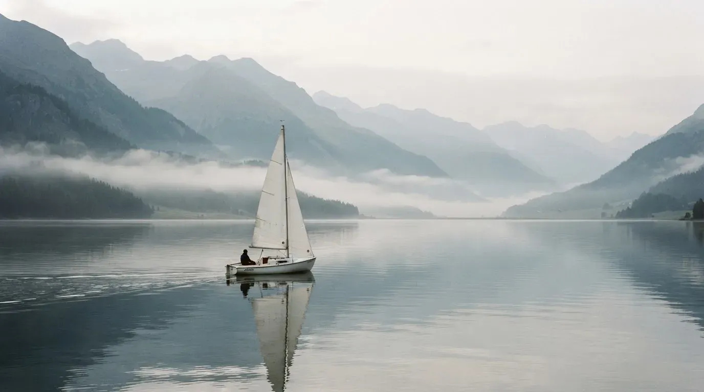 Ein kleines Segelboot auf einem ruhigen Bergsee, Nebel liegt am Ufer, die Berge verschwinden im Dunst