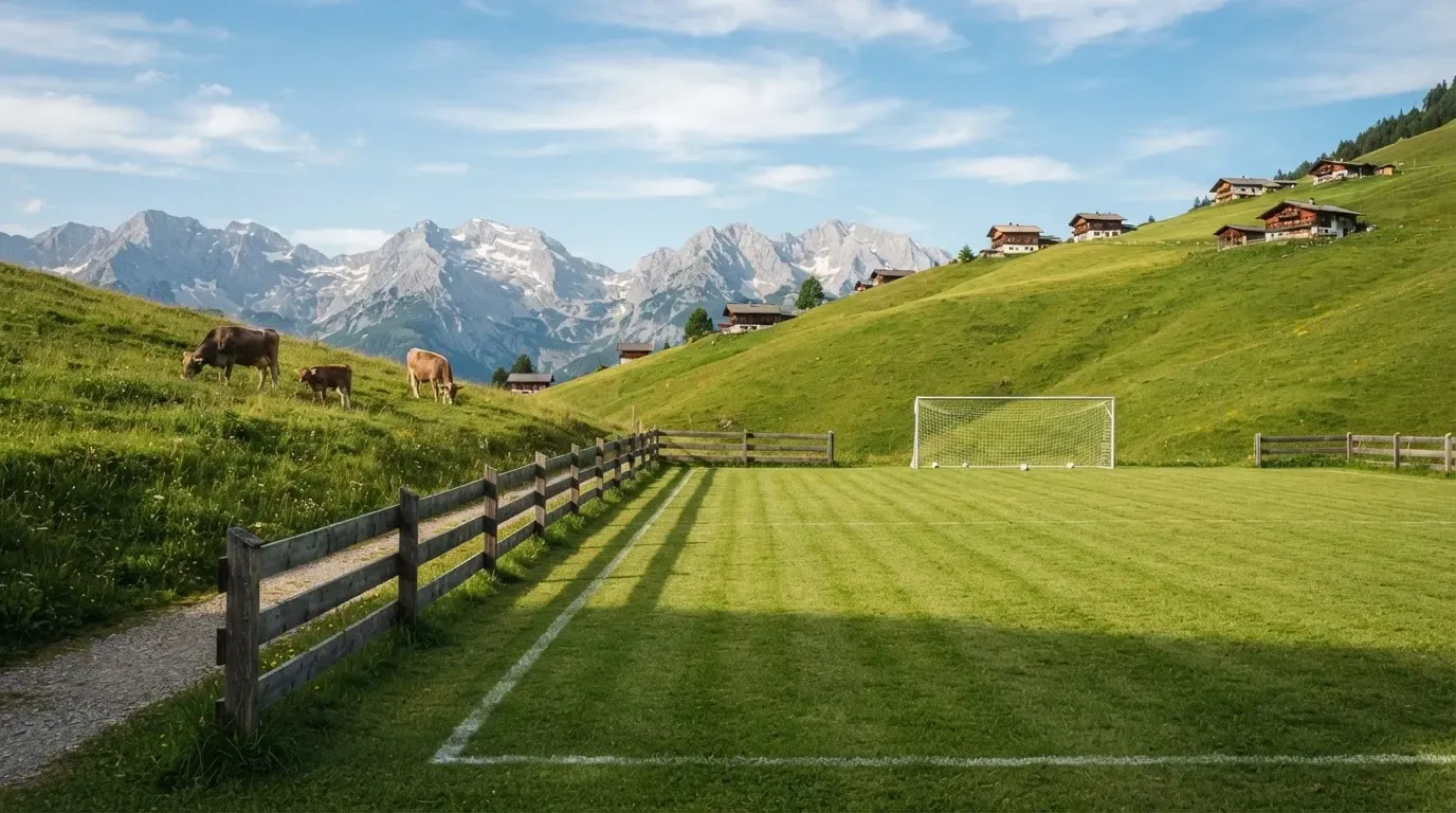 Ein Dorffussballplatz mit Holzzaun am Hang, dahinter grasen Kühe auf einer Weide unter hohen Wolken.