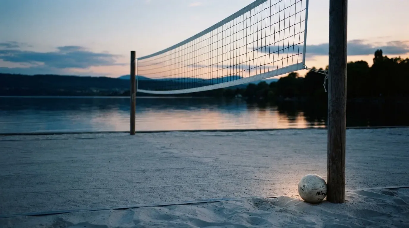 Ein Volleyballnetz steht an einem Seeufer in der Abenddämmerung, der Sand ist leer und das Wasser glatt.