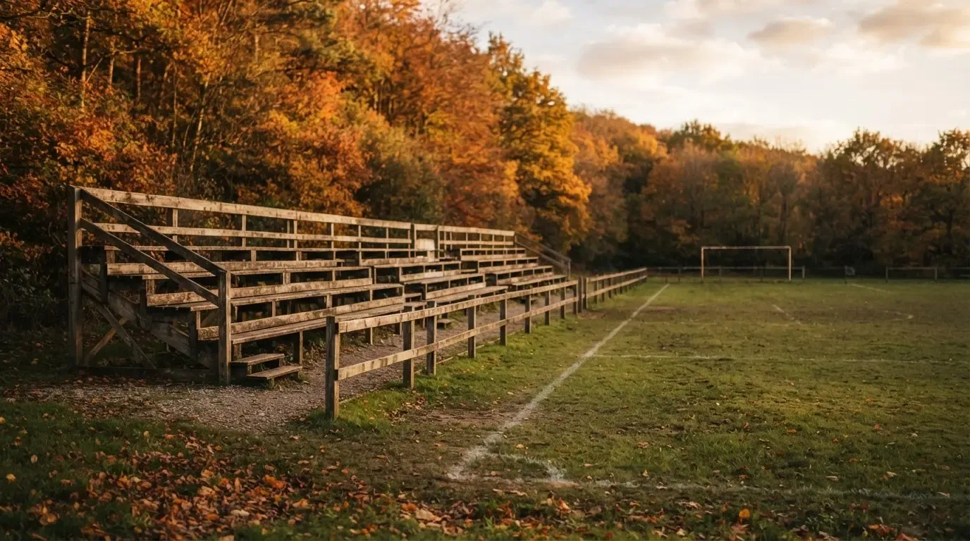 Eine leere Holztribüne steht am Waldrand neben einem kleinen Sportplatz im Herbstlicht.