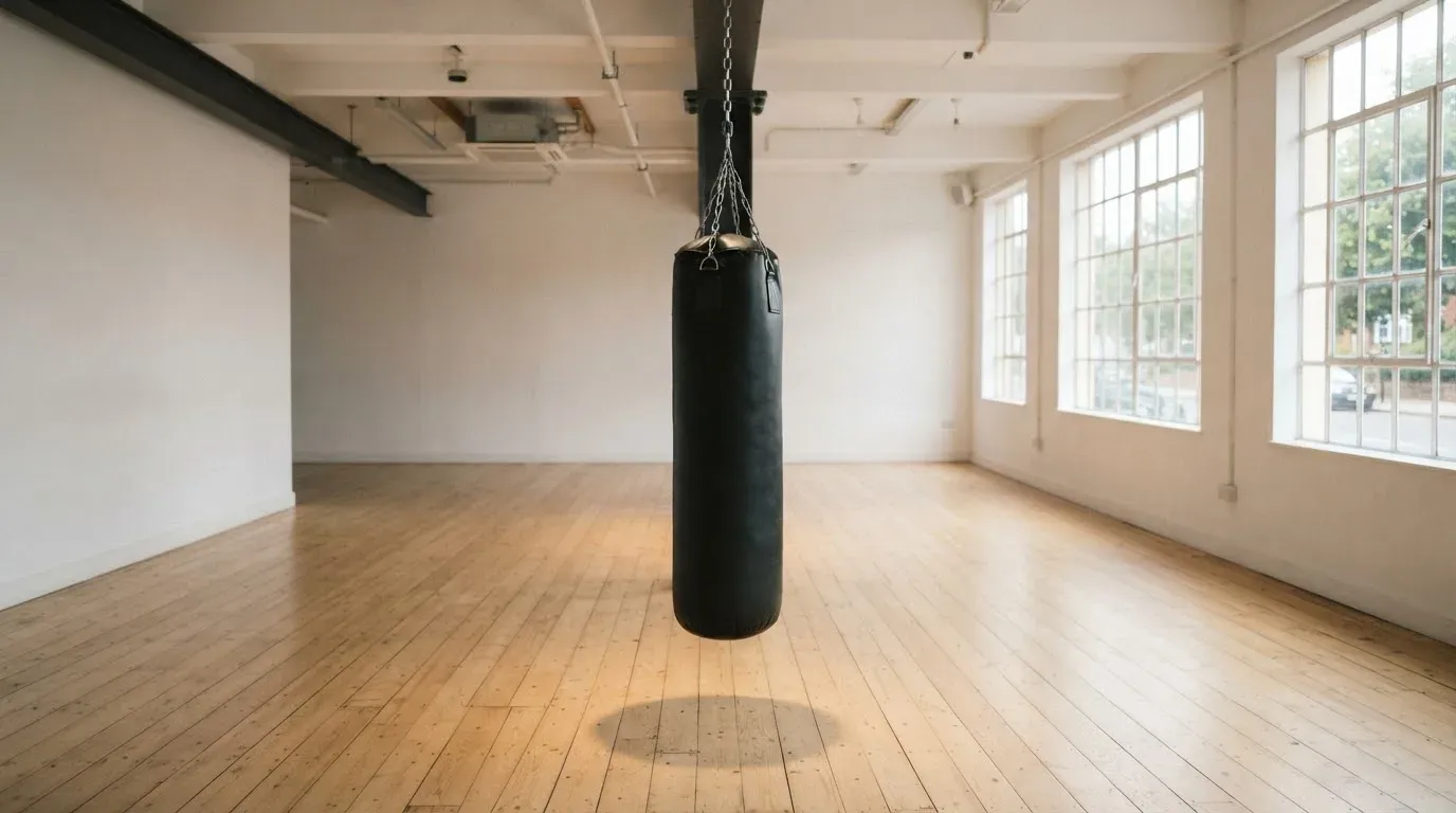 Ein Boxsack hängt an einer Kette in einem leeren Trainingsraum mit hellem Holzboden und grossen Fenstern.