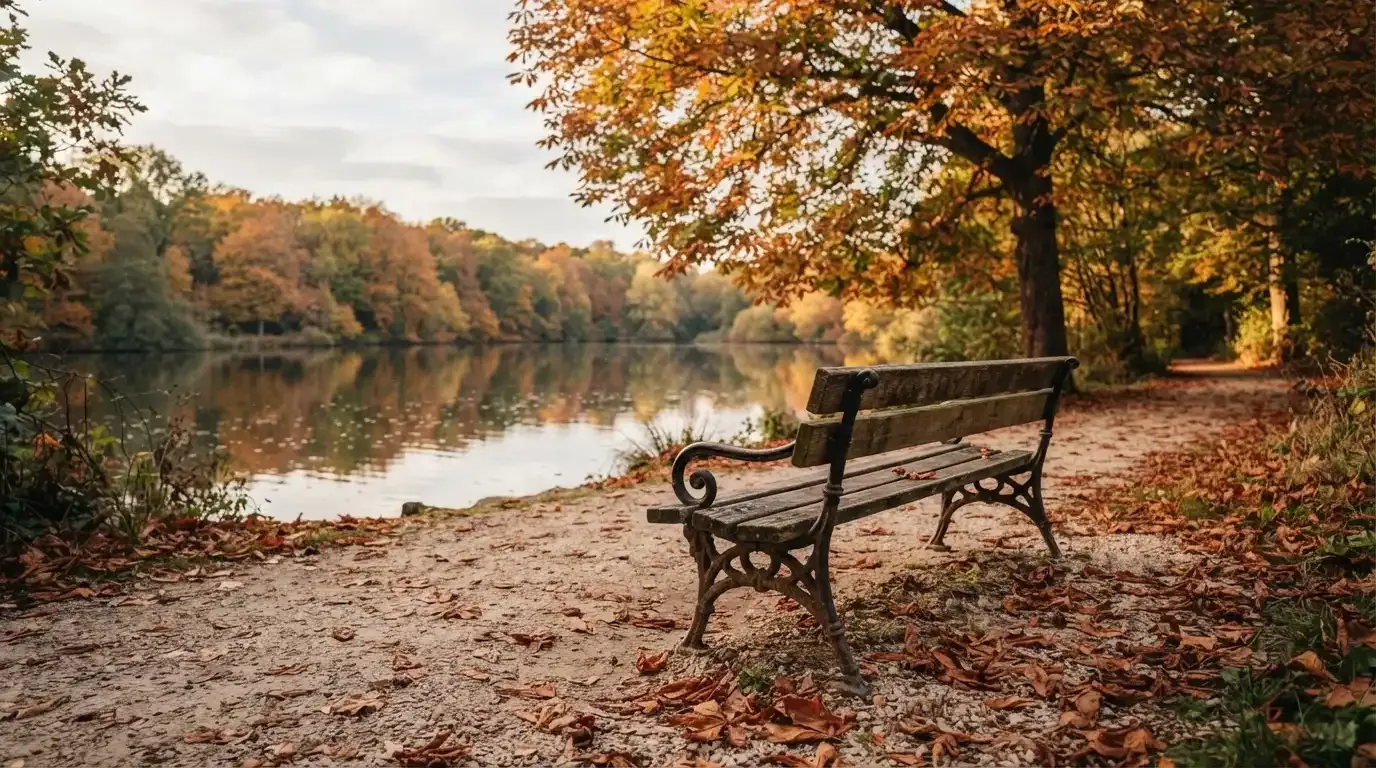 Eine Parkbank unter einer Kastanie, Herbstlaub bedeckt den Kiesweg, im Hintergrund ein stiller Teich.