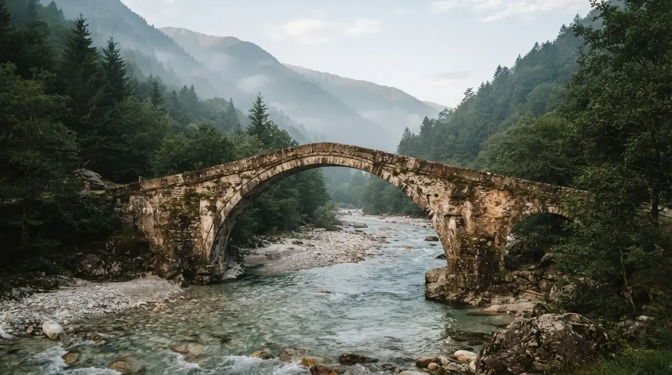 Eine alte Steinbrücke überspannt einen Gebirgsfluss, im Hintergrund steigen bewaldete Hänge an.